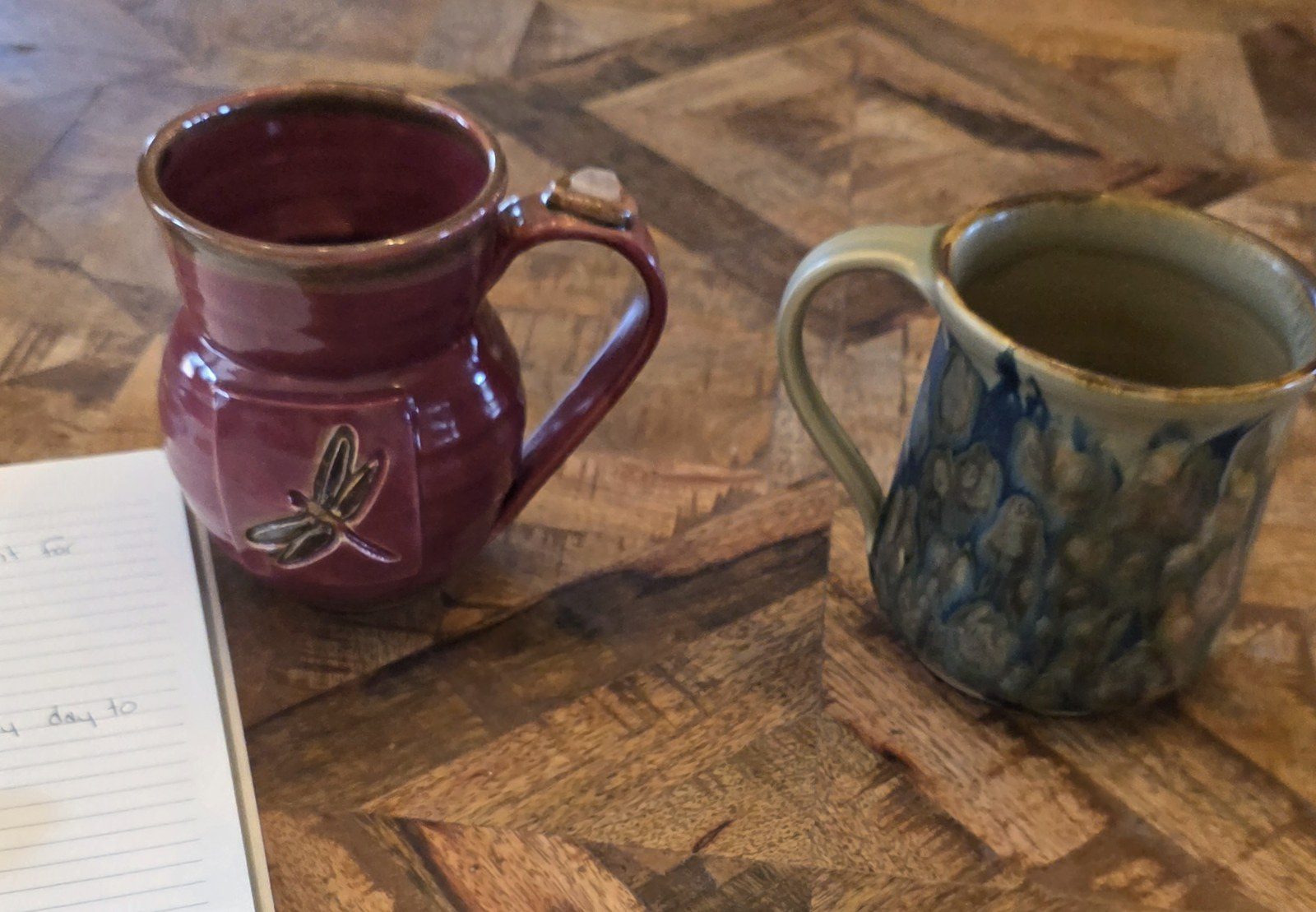 Two handmade ceramic mugs side by side on a wooden table next to a notebook of questions, a reminder not to face cancer appointments alone