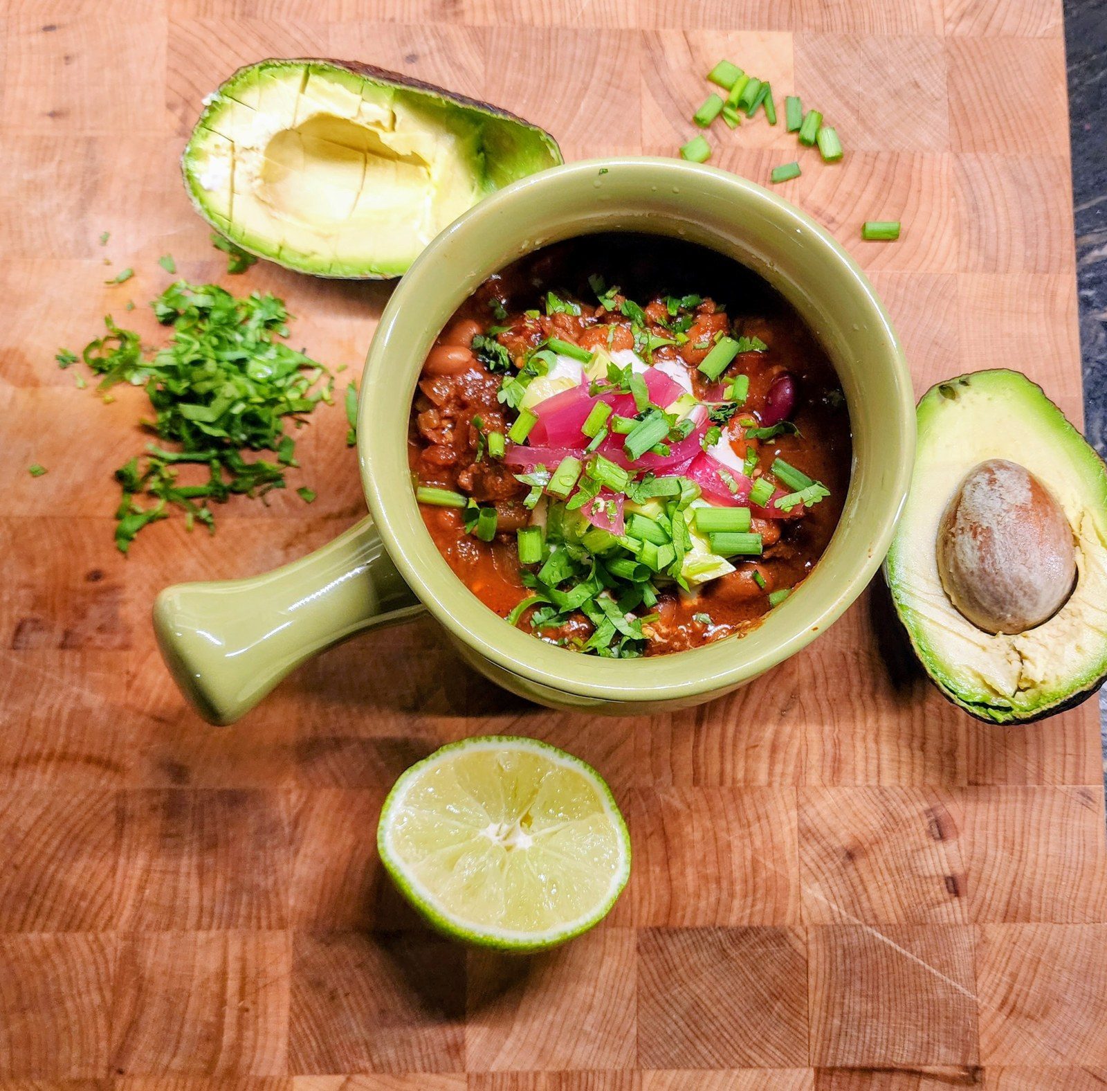 Homemade vegan chili in a green ceramic crock on a butcher block with avocado, lime and fresh herbs, warm comfort food and nourishment during cancer treatment