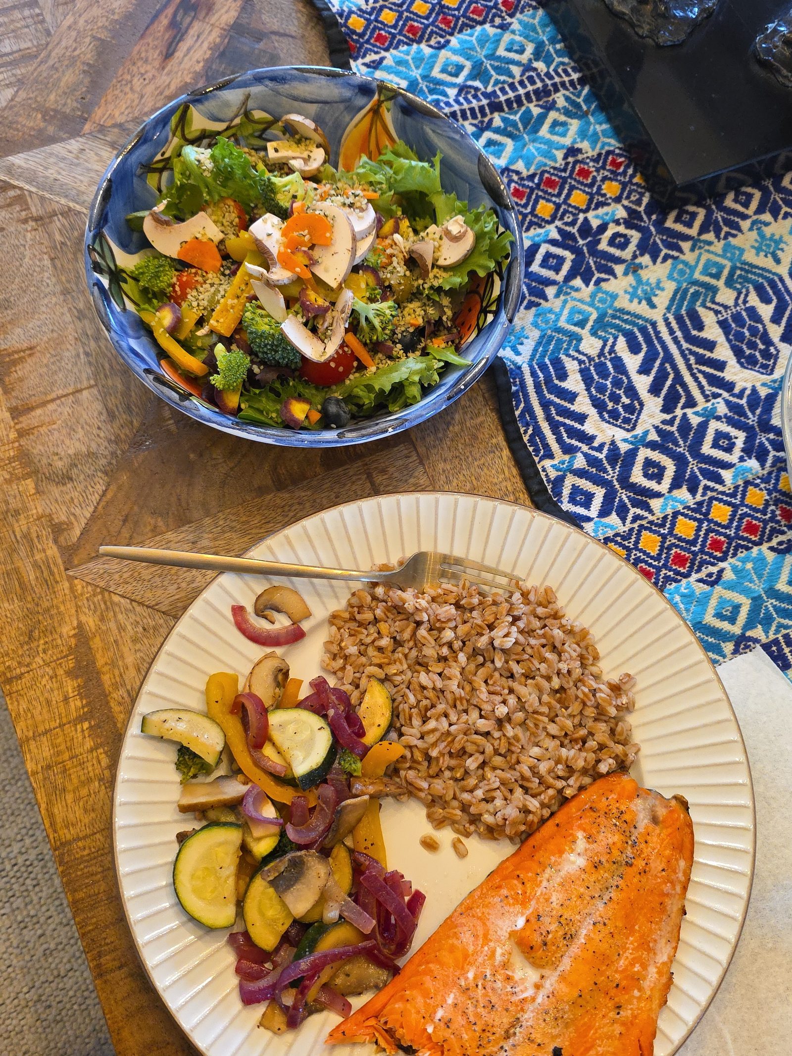 A plate of roasted salmon, farro, and sauteed zucchini, peppers and mushrooms beside a bowl of fresh green salad, set out for an unhurried meal