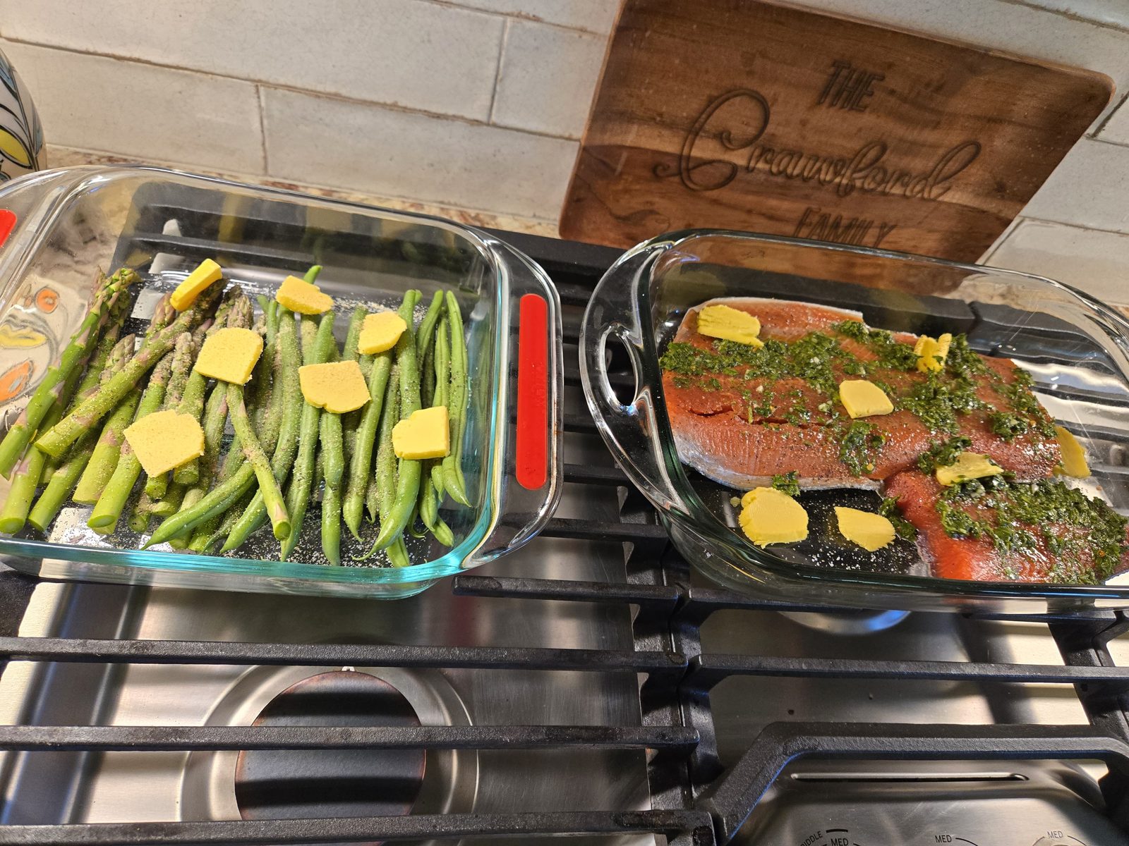 Salmon topped with herbs and butter beside asparagus and green beans in glass baking dishes on the stovetop, with a Crawford family cutting board behind