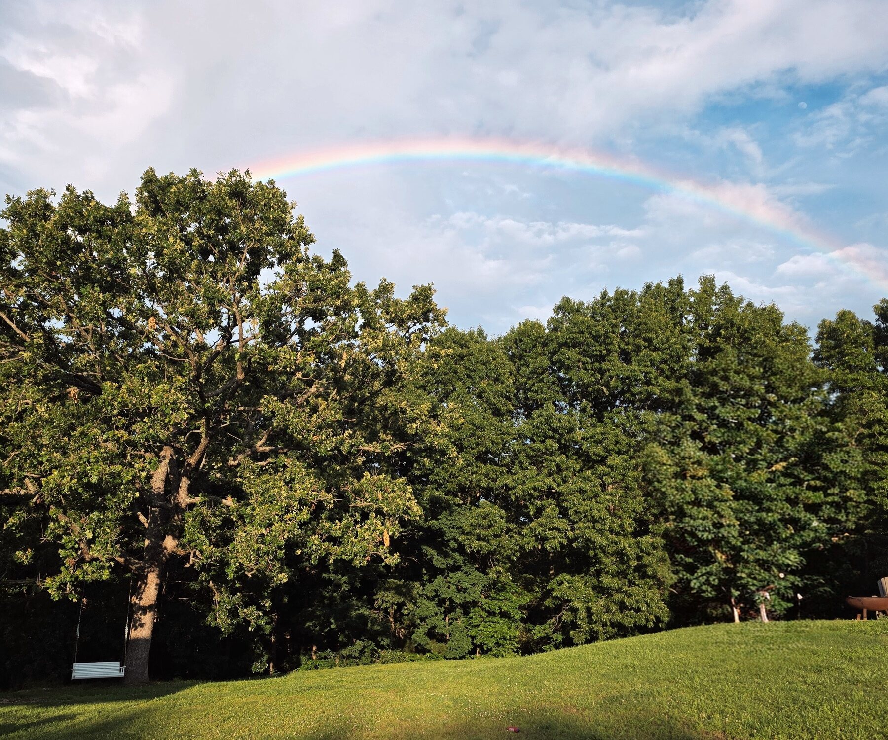 A rainbow arcing over a tree swing and oak tree on an Ozarks homestead at golden hour