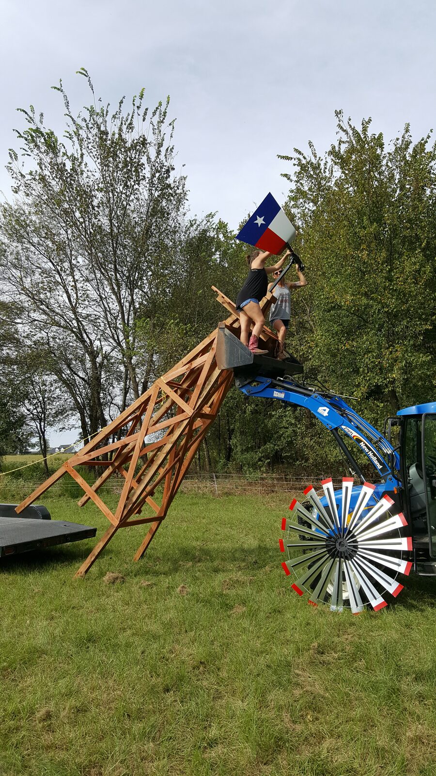 Two women on a tractor bucket lift building a windmill structure with a Texas flag held high