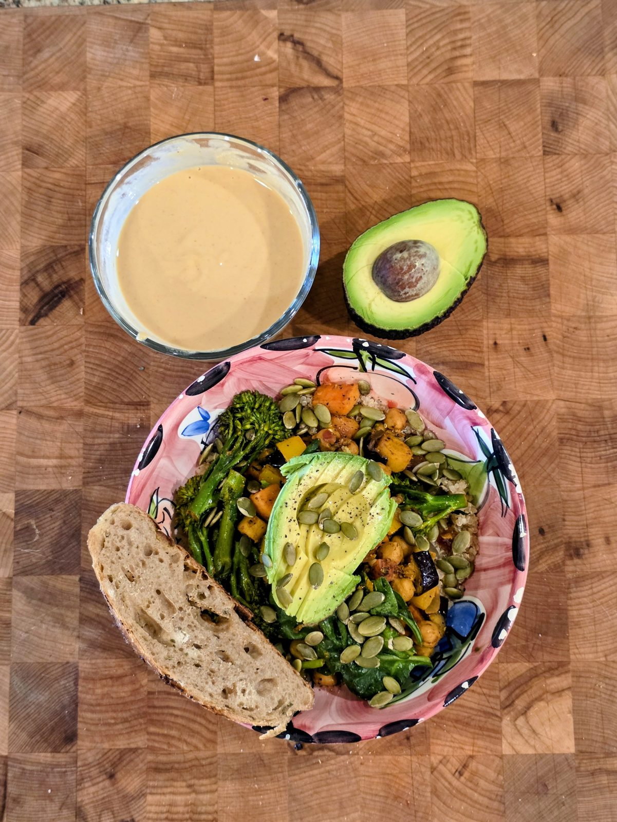 Whole foods bowl of quinoa, sautéed sweet potato, chickpeas, broccolini, asparagus, spinach, avocado and pumpkin seeds with lemon hummus dressing and a slice of homemade sourdough on a butcher block