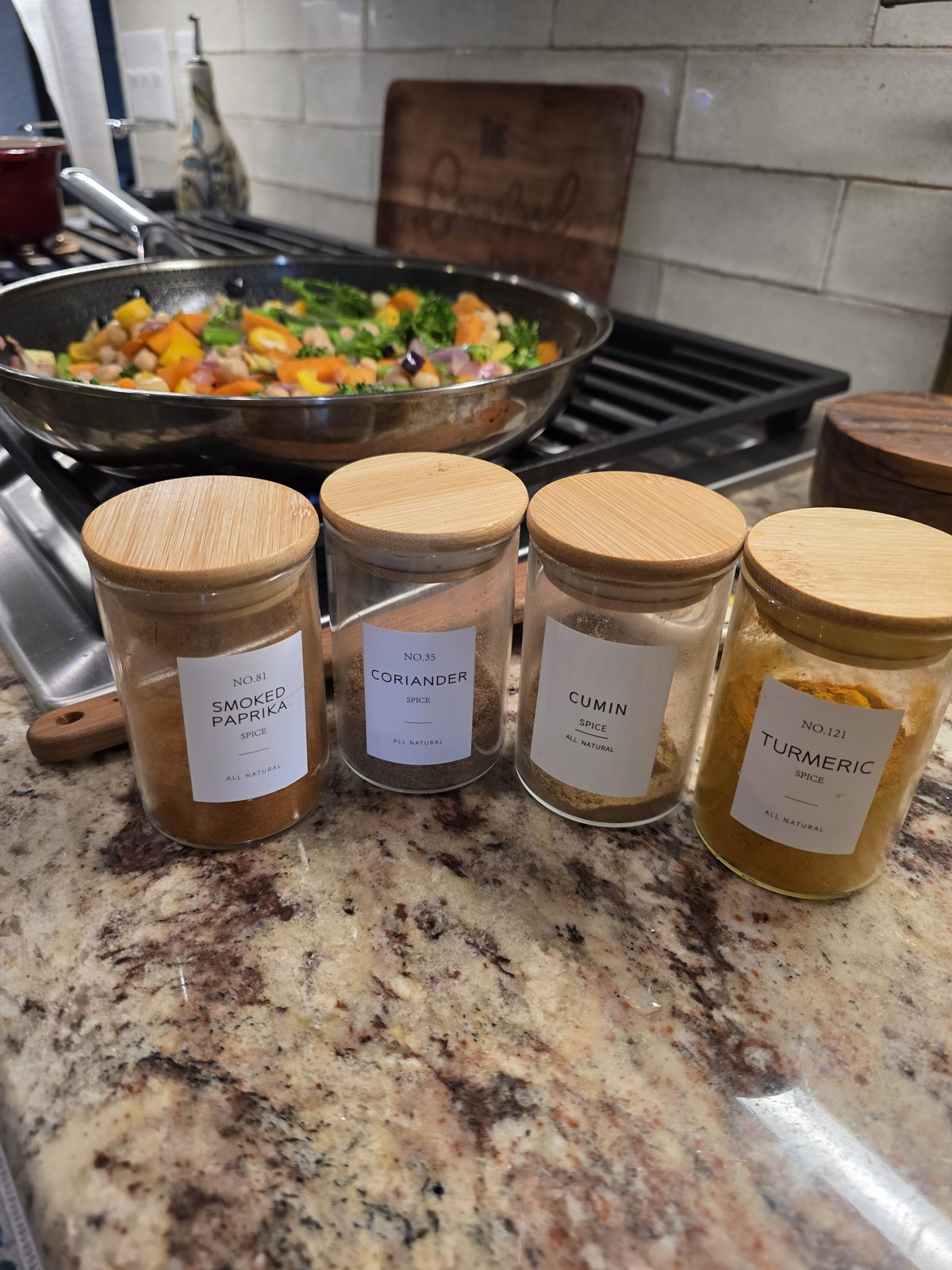 Glass jars of smoked paprika, coriander, cumin and turmeric beside a skillet of vegetables and chickpeas