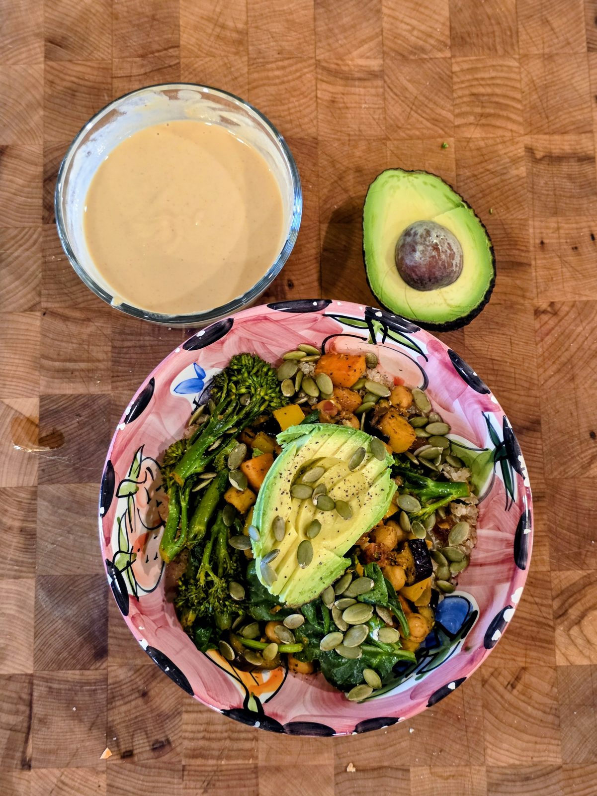 A colorful quinoa bowl with sautéed sweet potato, chickpeas, broccolini, spinach, sliced avocado and pumpkin seeds beside a bowl of lemon-sriracha hummus dressing
