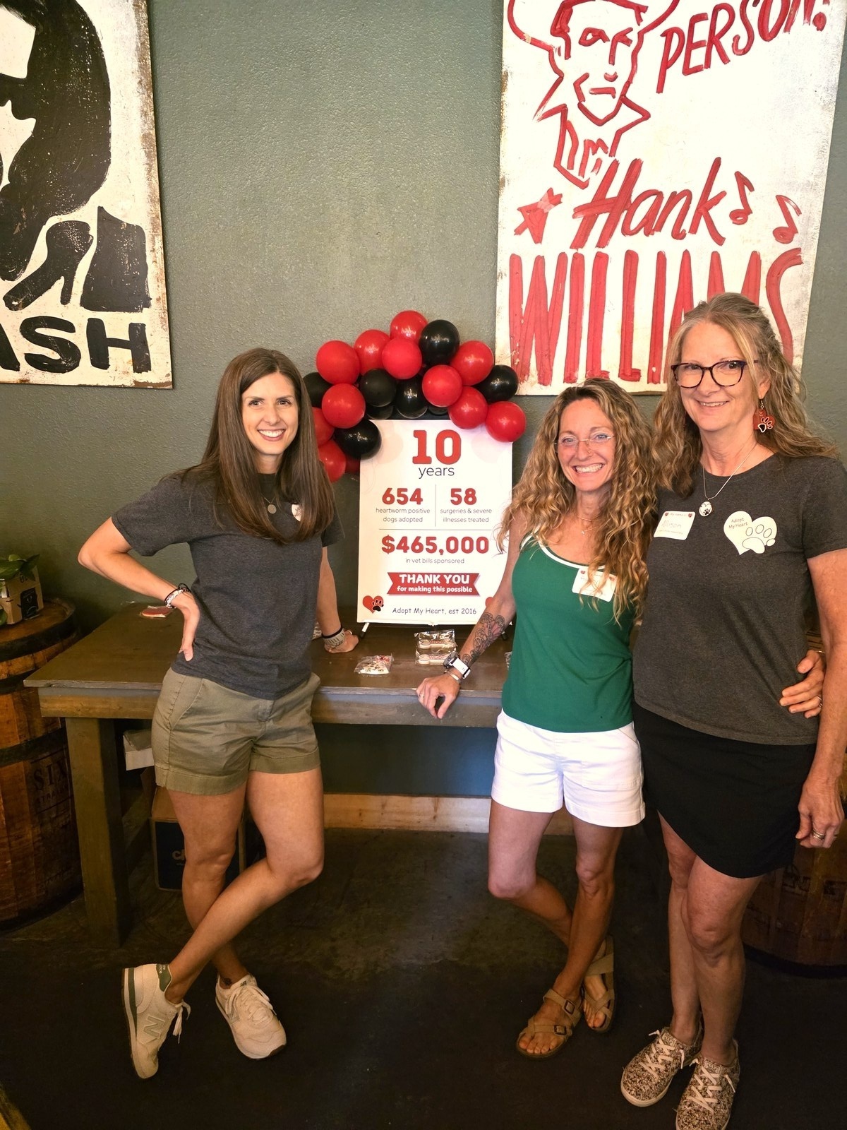 Three women celebrating Adopt My Heart's 10th anniversary in front of the stats sign at a Hank Williams venue in Texas