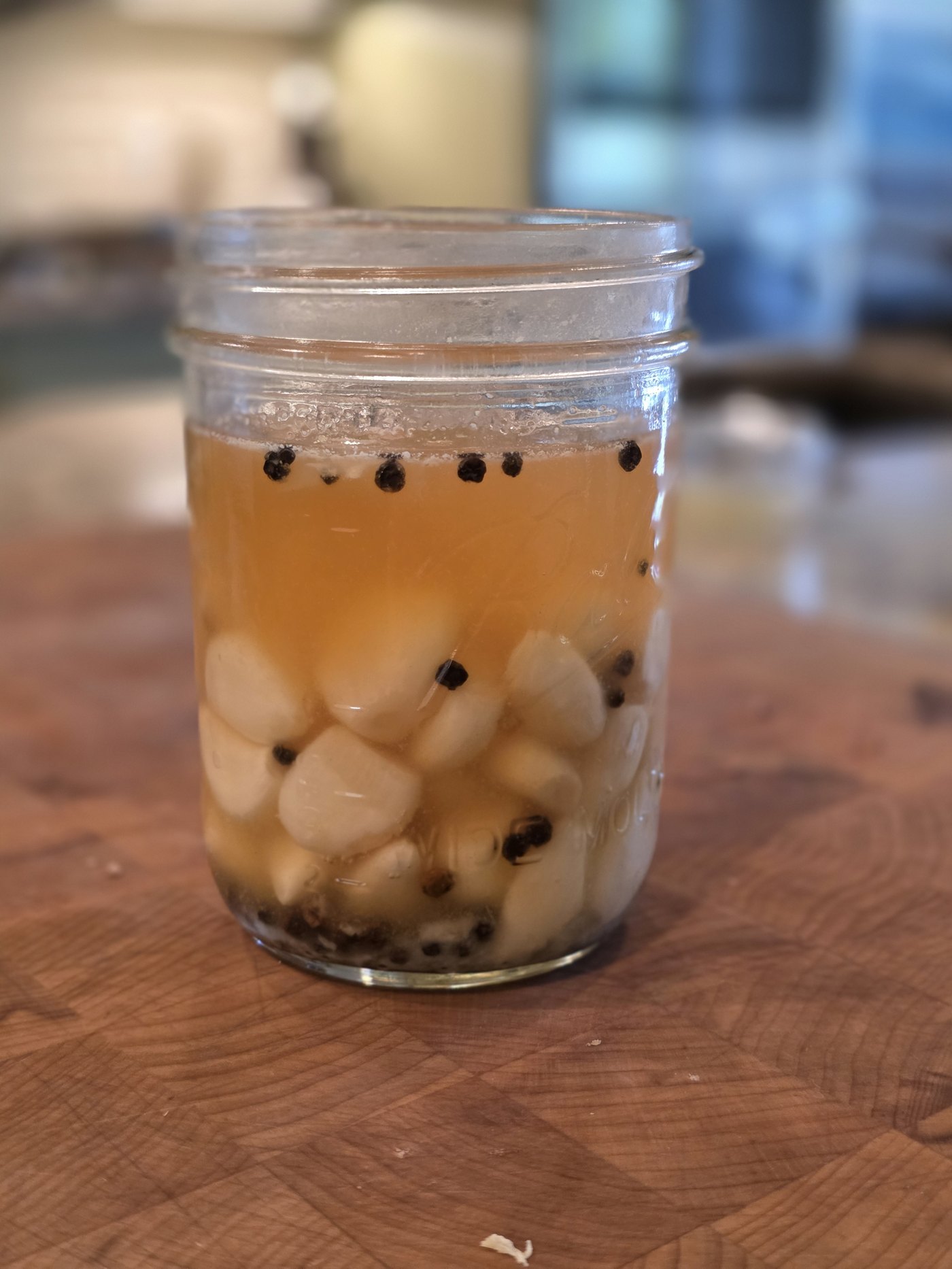 A close-up jar of lacto-fermented garlic cloves in golden brine with black peppercorns on a wooden board