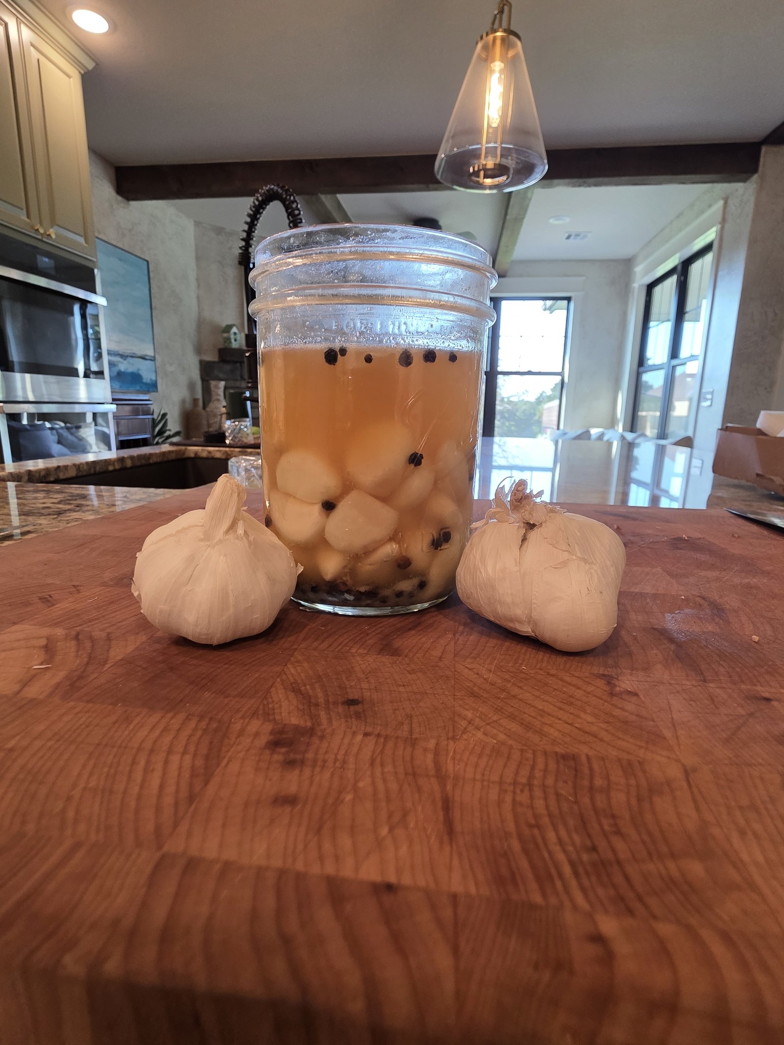 A jar of lacto-fermented garlic cloves in golden brine with black peppercorns, flanked by two garlic bulbs on a wooden board